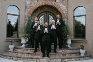 groom and groomsmen standing on steps, wearing black tuxedos, hands on the lapels of their coat