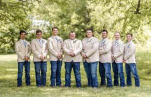 groom and groomsmen standing in a line, wearing tan jackets and blue jeans, hands held in front of them