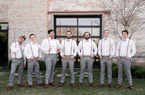 groom and groomsmen standing in a line, wearing light grey pants, white formal shirt, lavender bow ties and brown suspenders and shoes