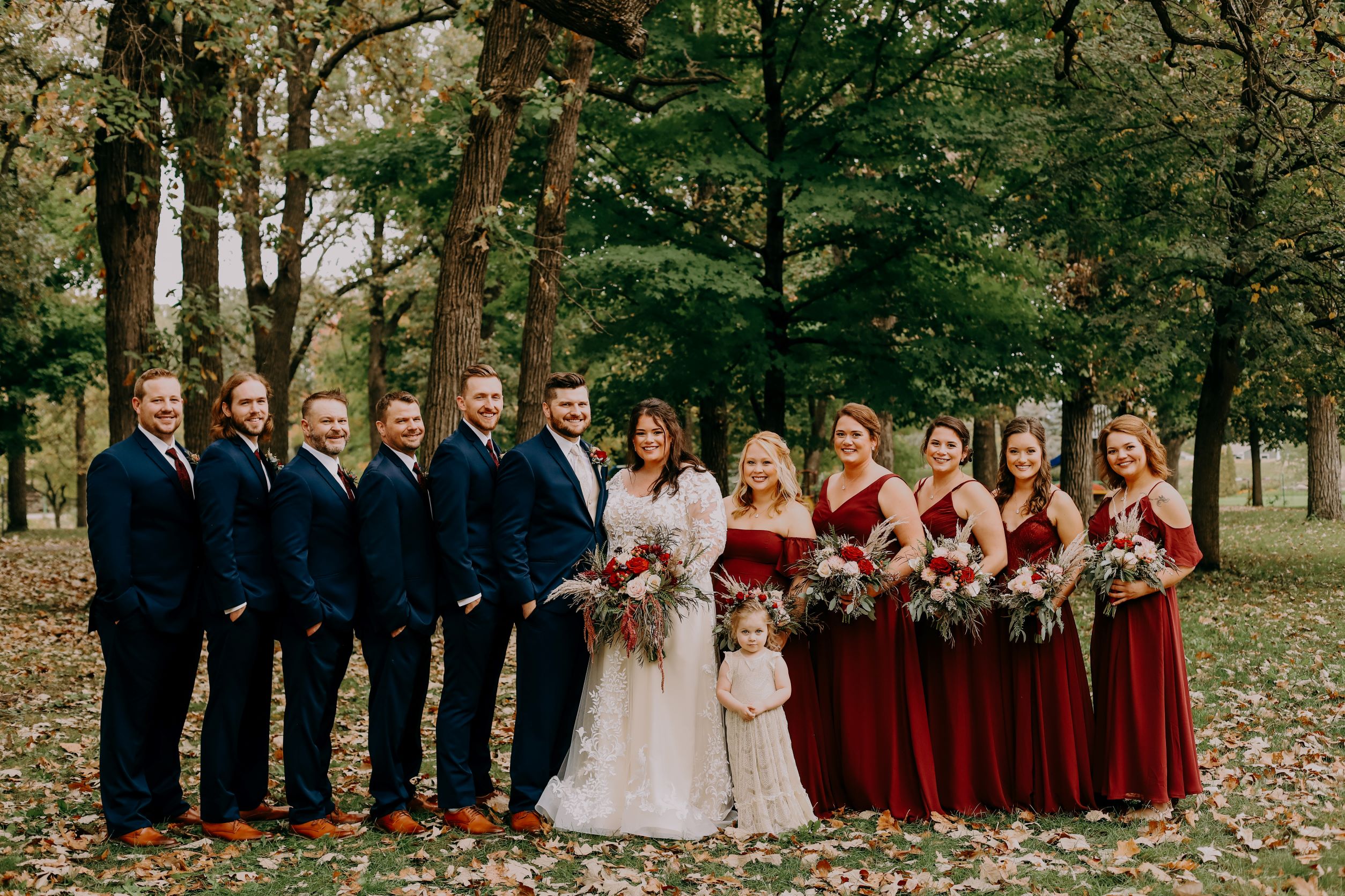 wedding party standing in line with trees in the background. guys in navy suits, ladies in deep red dresses