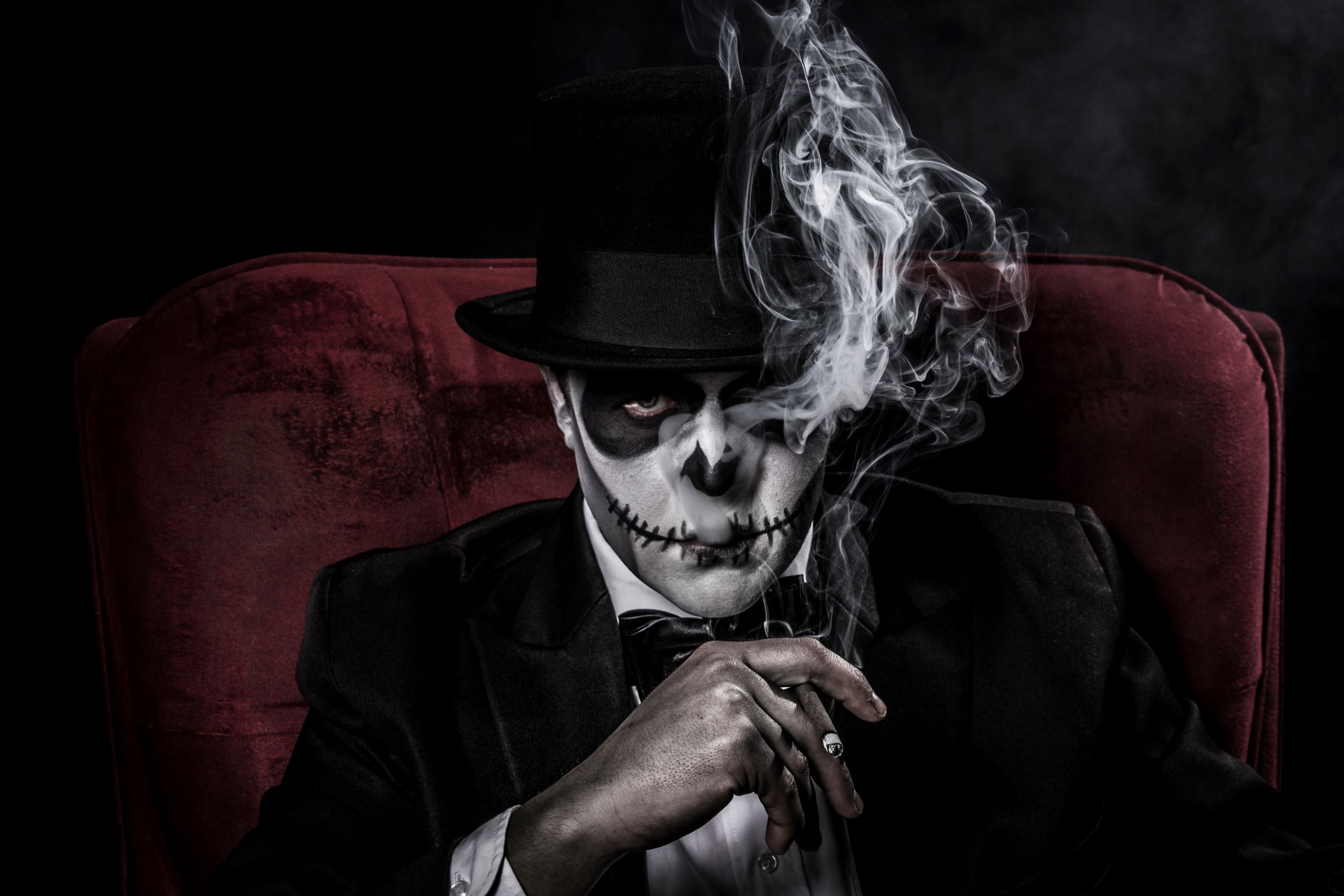 guy in black velvet coat and top hat sitting in red velvet chair, smoking a cigar, smoke coming out of his mouth, face painted like a skeleton