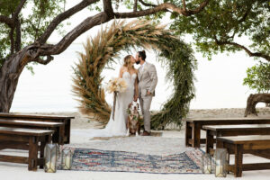 Bride and groom on a beach, standing in front of a large greenery wreath, kissing. Dog is sitting in front of them