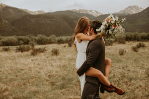 bride and groom at their destination wedding in field with mountains in the background. groom holding up bride and bride has legs wrapped around groom