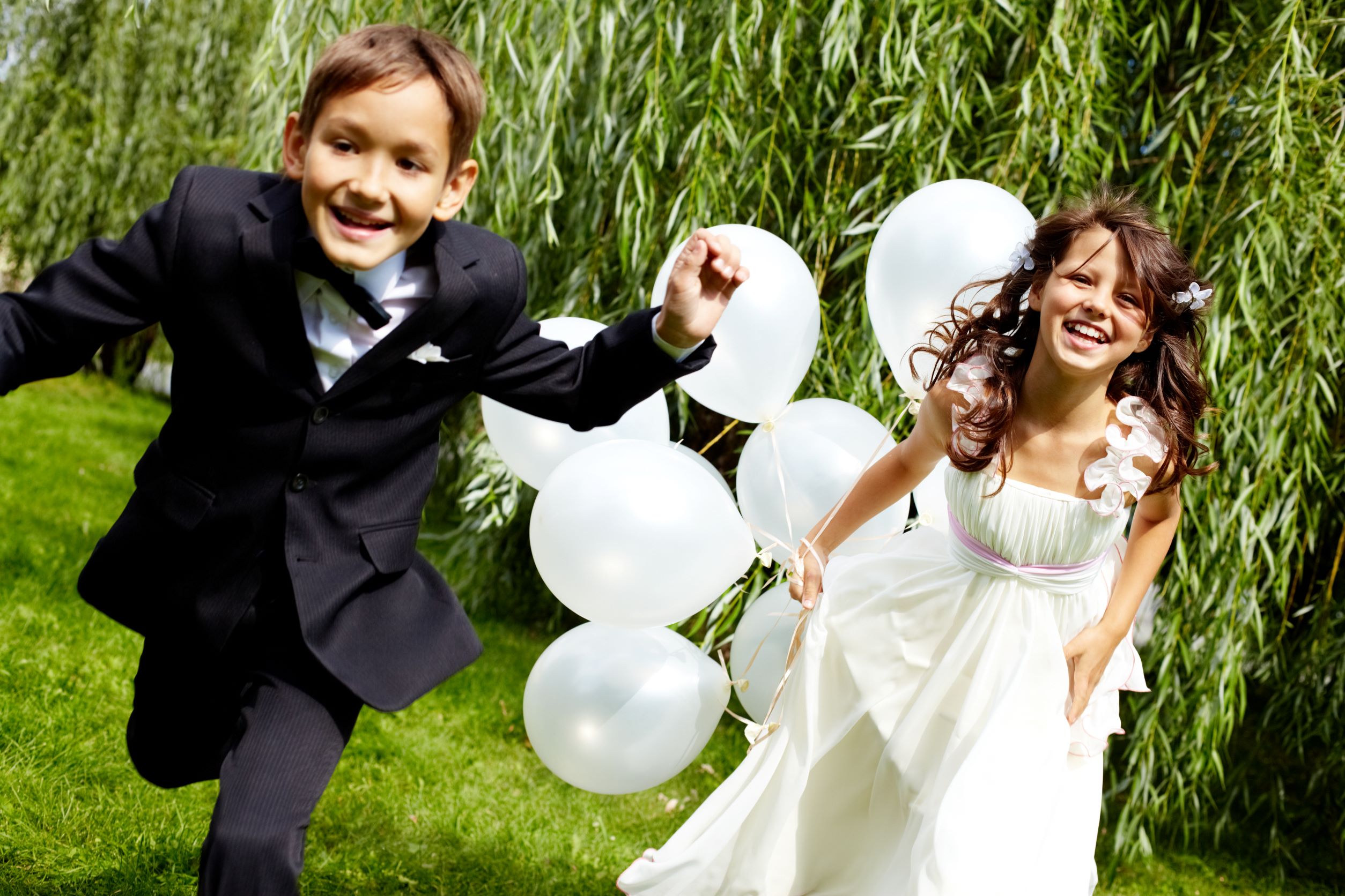 little boy in black tux being chased by little girl in flower dress holding white balloons
