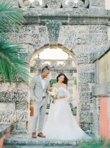 groom in tan suit, no tie, looking at his bride, both standing in front of an Aztec-styled archway 