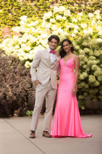 guy in tan suit with bright pink bow tie, standing next to his date. She is wearing a hot pink dress. A flower wall is behind them.