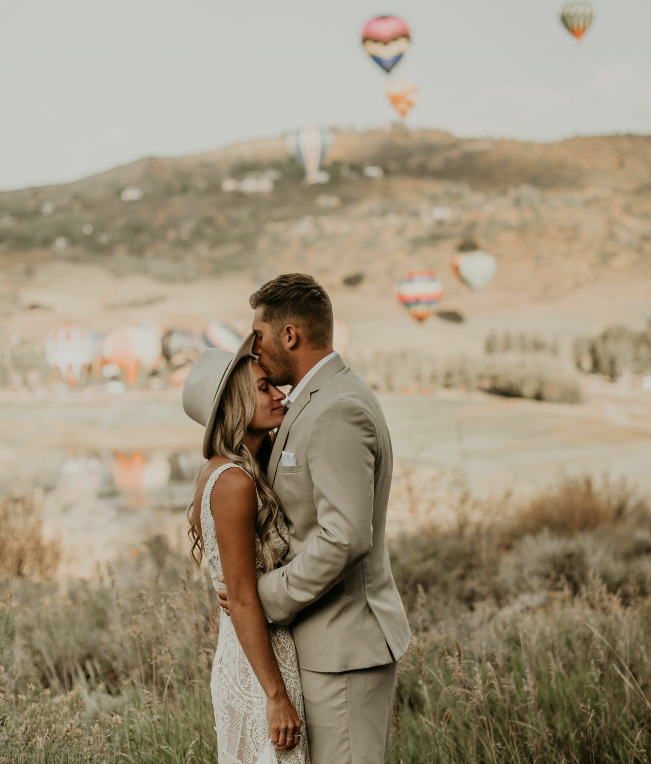 bride getting kissed on the forehead by groom who is wearing tan suit. Background has hot air balloons