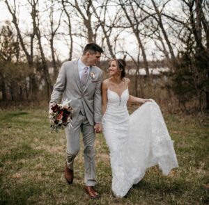 bride and groom walking in field, hand in hand. Groom in tan suit, holding bouquet