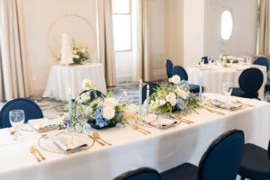 table settings with white tablecloths, navy chairs, white flowers, and gold flatware