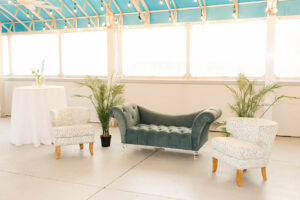 seating area for a destination wedding including a blue couch, white cushioned chairs, ferns, and a bar height table