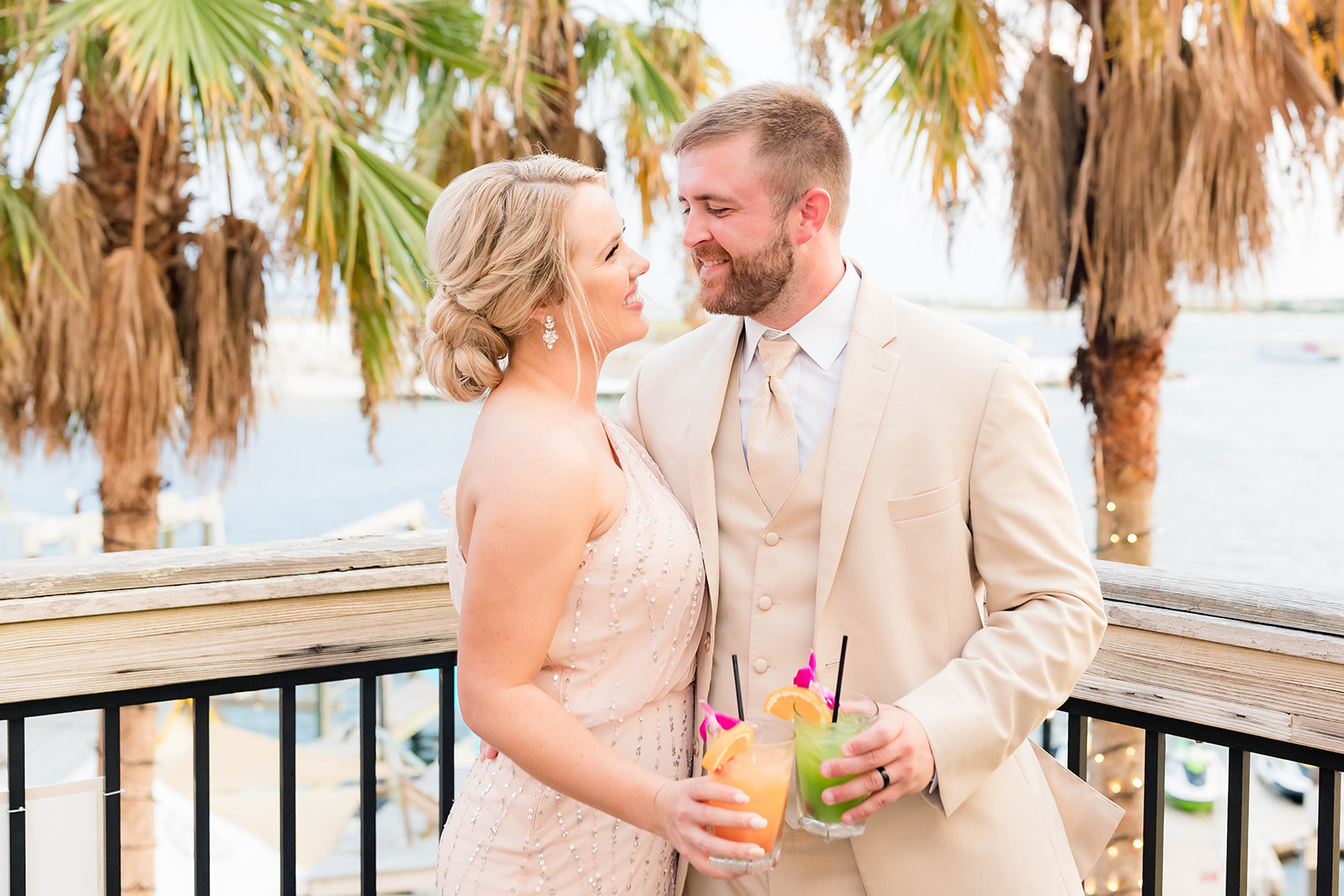 bride and groom toasting with colorful drinks on a patio overlooking a beach and water inlet