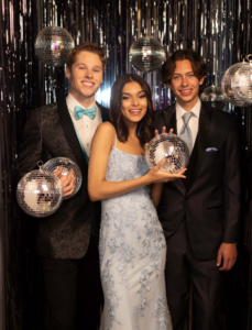two high schoolers in tuxedos standing close to a girl in a blue homecoming dress