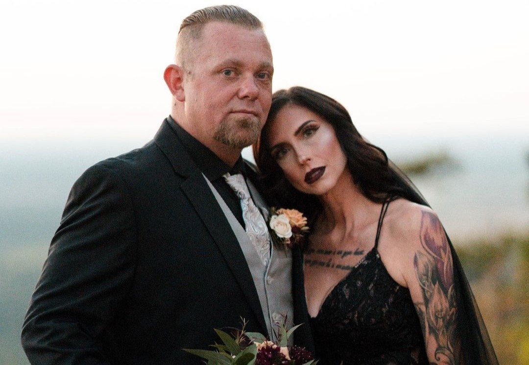 Bride in black wedding dress, her head is on the groom's shoulder. Groom in black tux, silver vest and tie, holding moody wedding bouquet