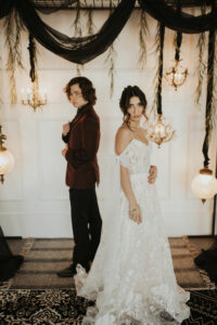 bride in white dress looking at camera, groom in burgundy coat, black pants standing behind her to the left. Black drapes and low hanging chandeliers around them. 