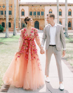 Valentine's Day look: lady in floral dress in shades of pink, coral and red and the man in a a tan suit, white button down shirt and white loafers