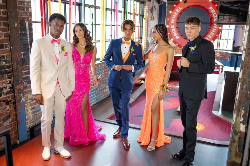 group of prom kids wearing tuxedos, suits, and long dresses in neon or bright colors at an indoor mini-golf location