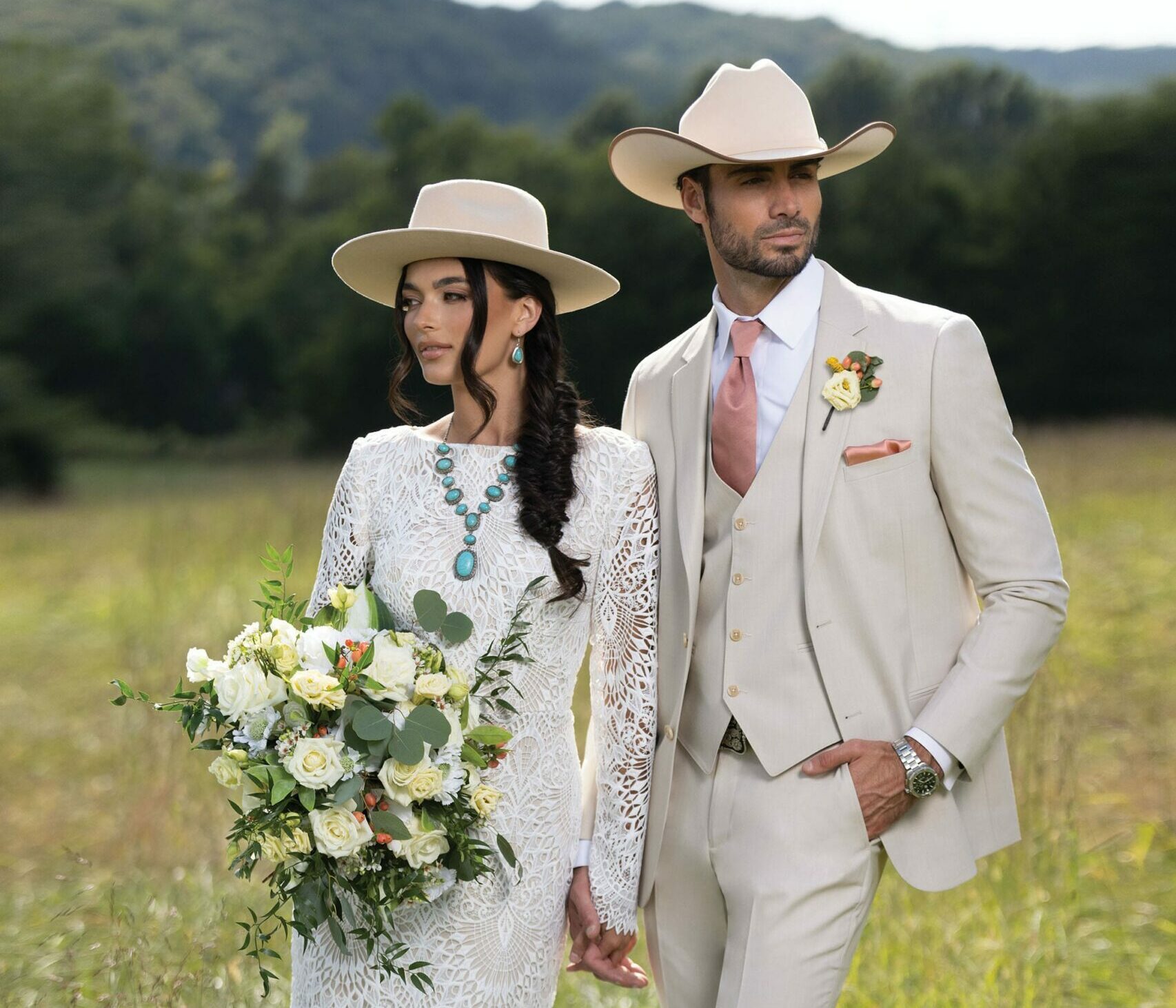 bride in a boho look with white lace dress, turquoise jewelry, and brimmed hat standing with the groom in a Tan Michael Kors Suit and western hat