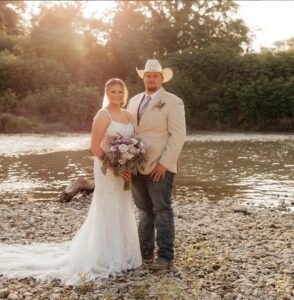 bride and groom wearing a tan jacket, blue jeans, and a western cowboy hat in front of a river
